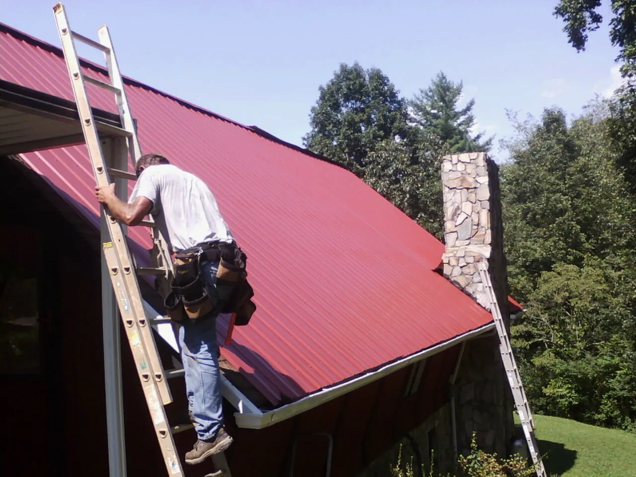 Roofer on a ladder beside a red metal roof and stone chimney — Soffit & Fascia Repair by Roofing Brothers in Paint Lick